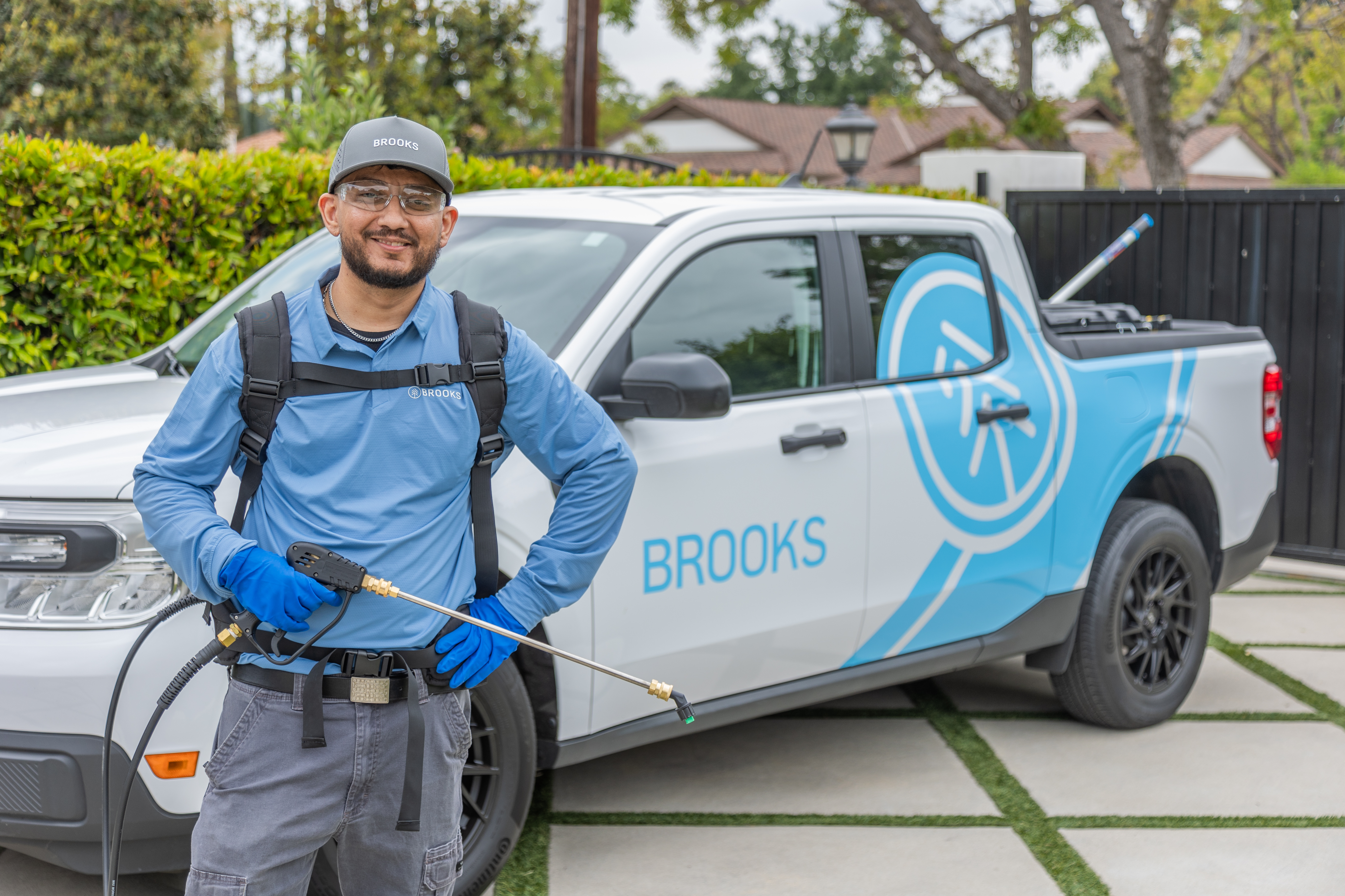Brooks Pest Control technician with sprayer in front of branded truck in suburban driveway